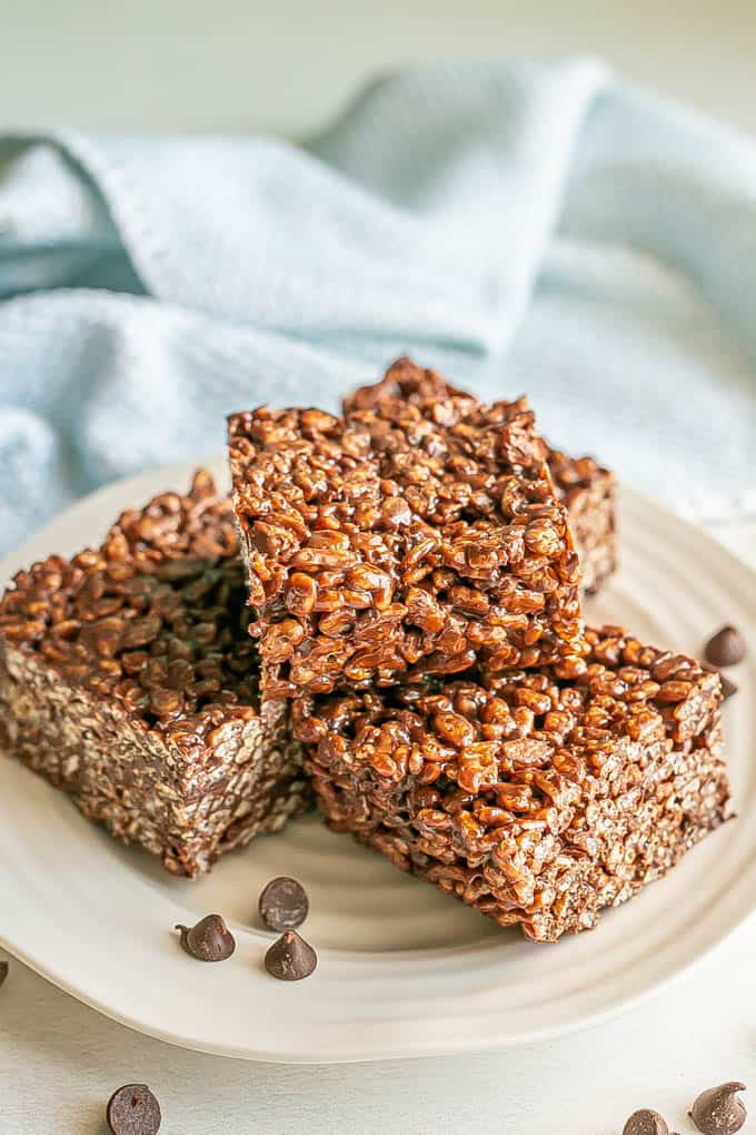 A stack of chocolate rice krispie treats on a white plate with chocolate chips scattered nearby