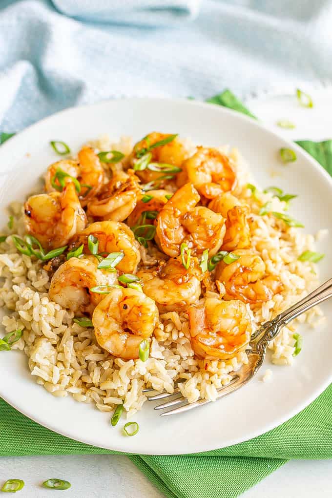 Honey garlic shrimp piled over some steamed brown rice on a white plate, topped with sliced green onions with a fork resting on the plate