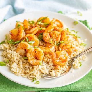Honey garlic shrimp piled over some steamed rice on a white plate, topped with sliced green onions with a fork resting on the plate