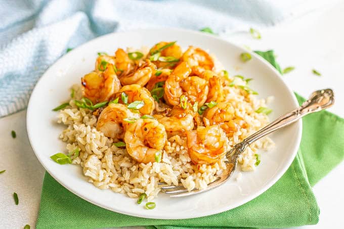 Honey garlic shrimp piled over some steamed rice on a white plate, topped with sliced green onions with a fork resting on the plate