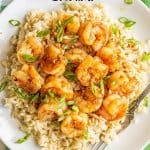 Honey garlic shrimp piled over some steamed brown rice on a white plate, topped with sliced green onions with a text overlay on the photo