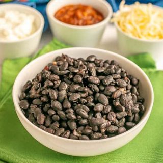 A white bowl of cooked black beans and bowls of toppings in the background