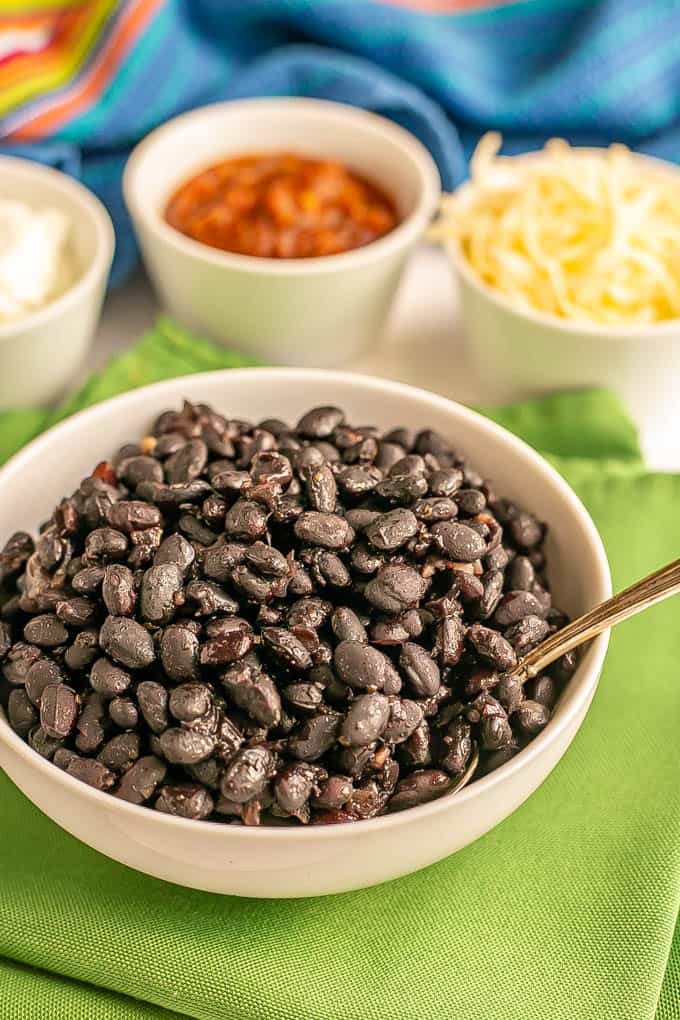 A white bowl of cooked black beans with a spoon resting in it and bowls of toppings in the background