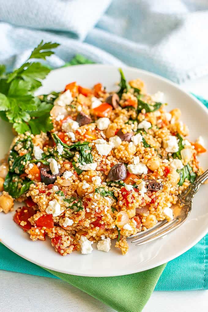 A white dinner plate of a vegetarian quinoa mix with chickpeas, spinach and tomatoes with a fork alongside and feta cheese and olives on top