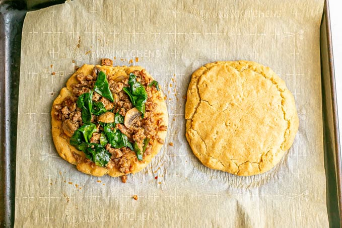 Two flatbreads on a parchment paper lined baking sheet, one topped with a sausage and veggie mixture