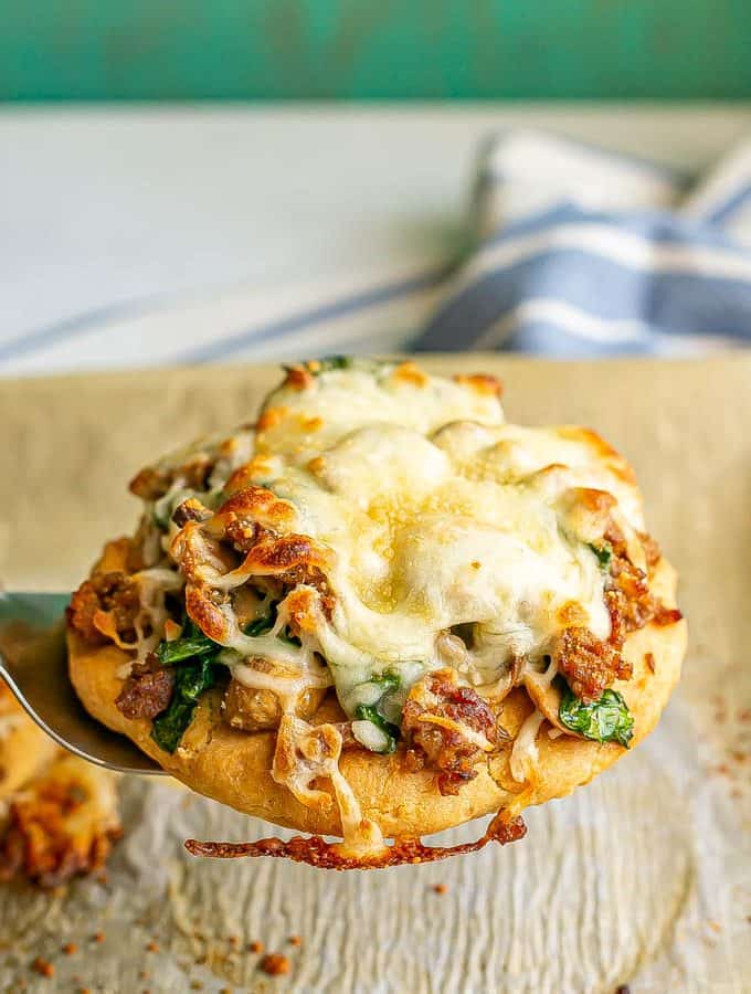 A spatula picking up a flatbread topped with sausage, mushrooms, spinach and cheese