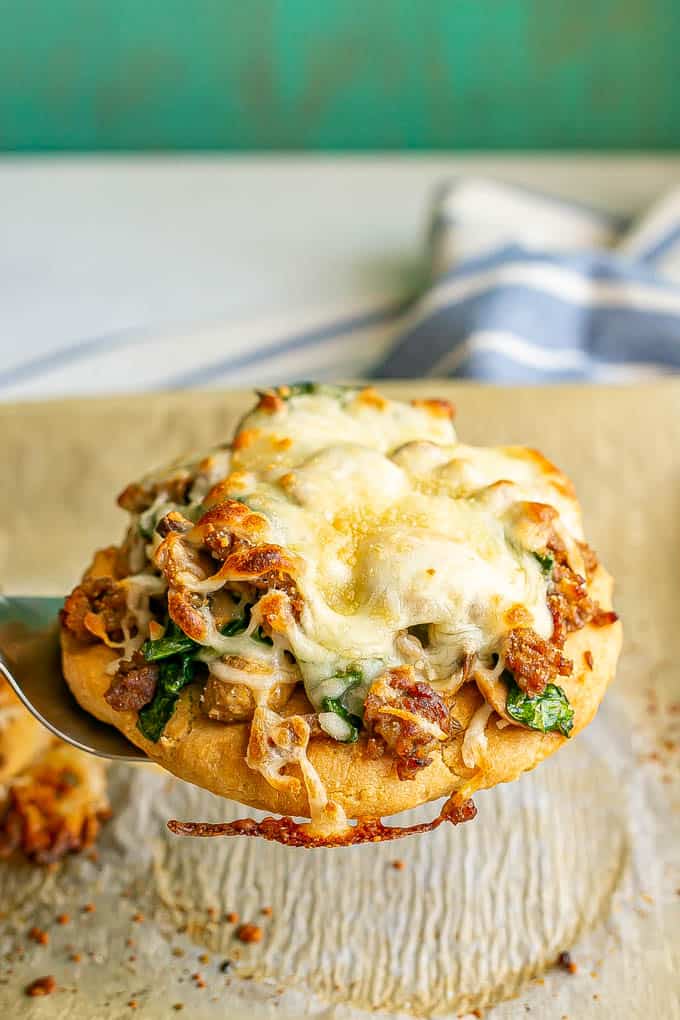 A spatula picking up a flatbread topped with sausage, mushrooms, spinach and cheese