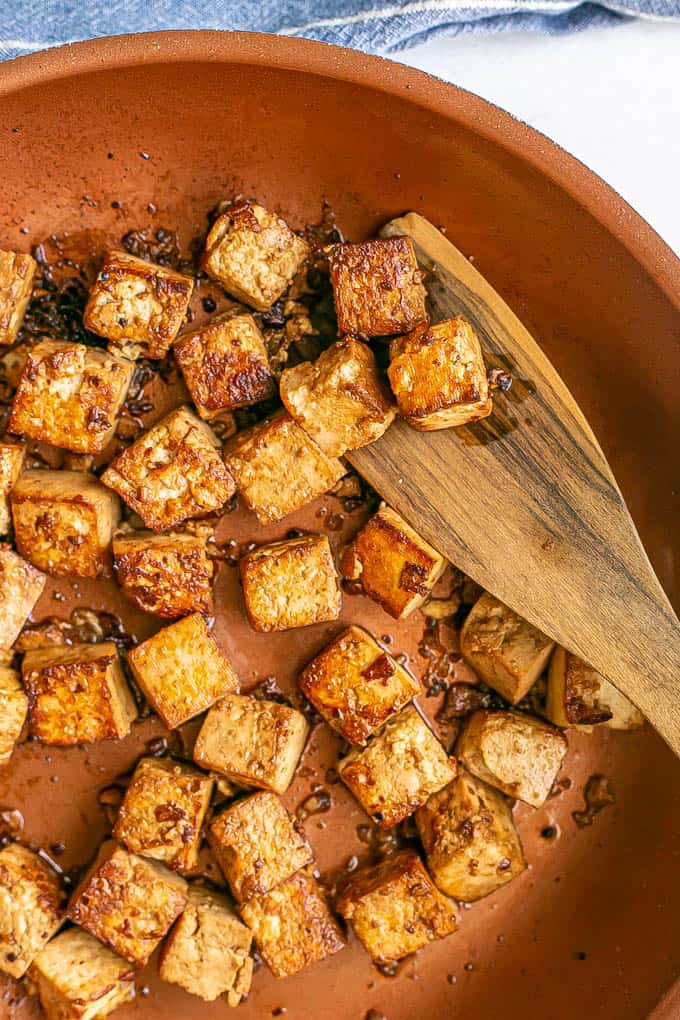 Crispy browned cubed tofu in a copper skillet with a wooden spatula scooping some up