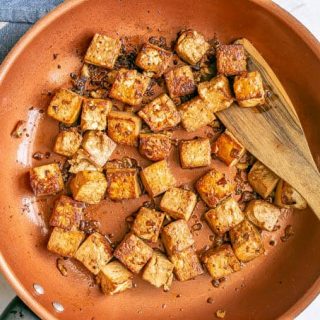 Crispy browned cubed tofu in a copper skillet with a wooden spatula scooping some up