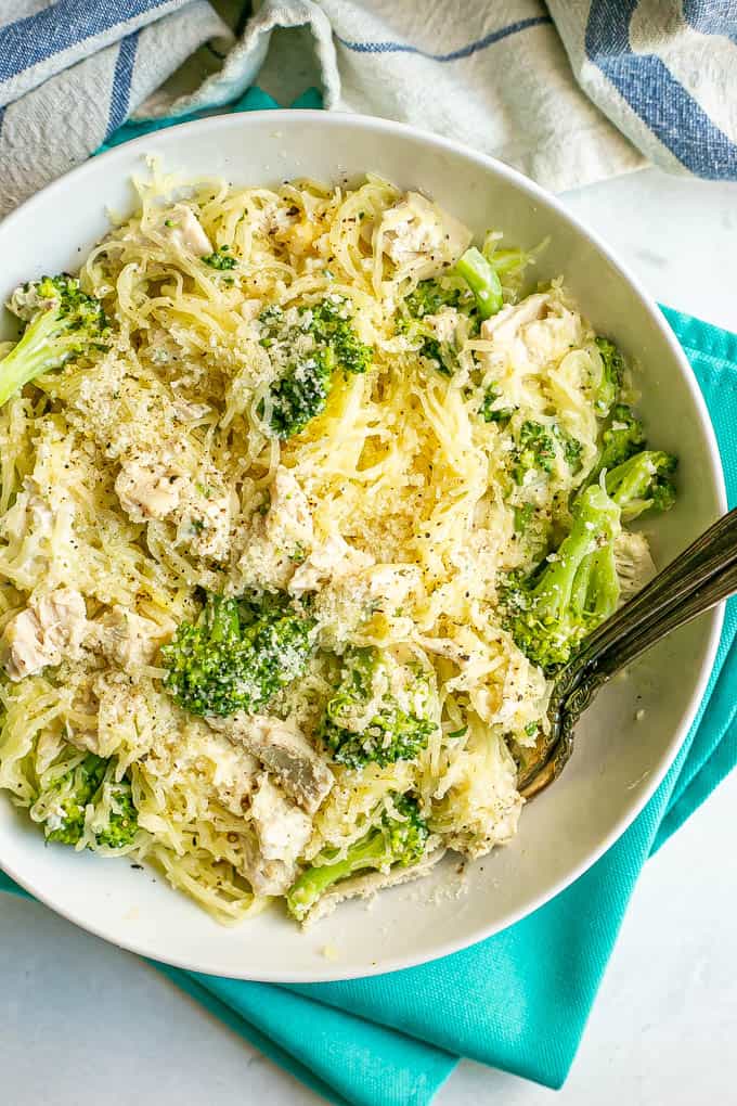 A white dinner bowl filled with spaghetti squash mixed with chicken and broccoli and topped with Parmesan cheese with two forks on the side of the bowl