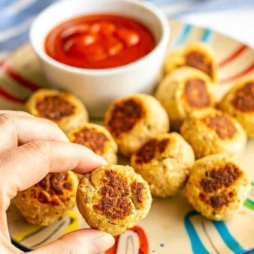 A hand picking up a small meatball from a plate of small cooked meatballs