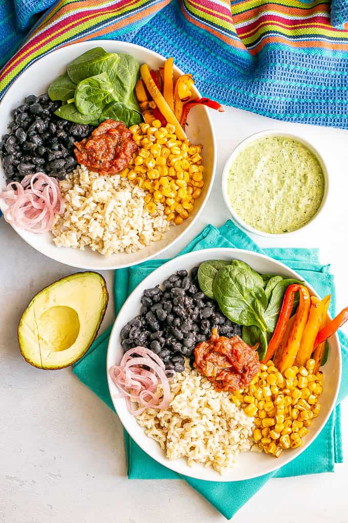 Two arranged bean and rice bowls with veggies and a half avocado and a bowl of green dressing nearby