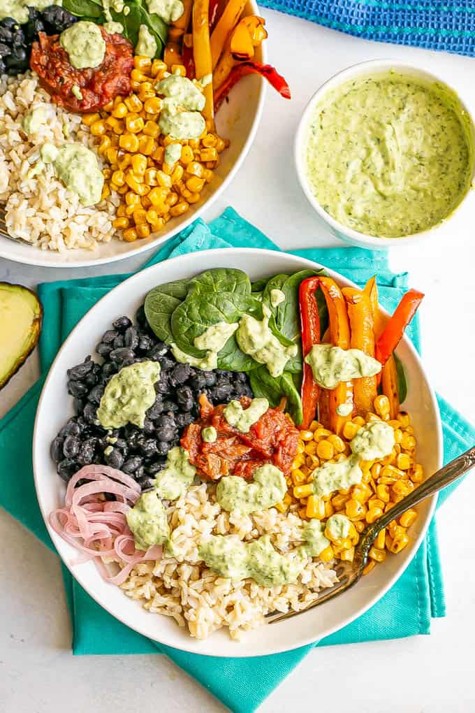 Two white bowls served with brown rice, black beans, spinach, peppers and corn, plus a cilantro lime dressing drizzled over top