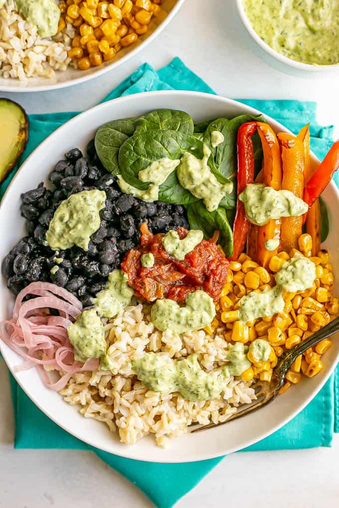 A large white bowl with brown rice, black beans, spinach, peppers and corn, plus a cilantro lime dressing drizzled over top