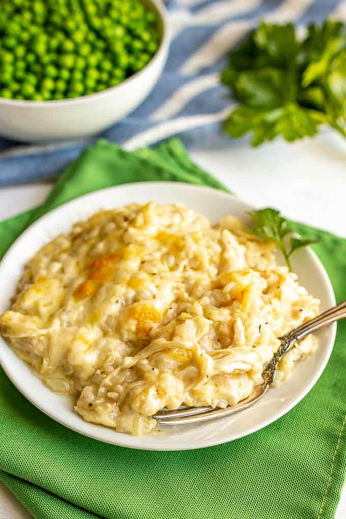 A cheesy chicken and rice mixture served on a small white dinner plate with a fork alongside and a bowl of green peas in the background