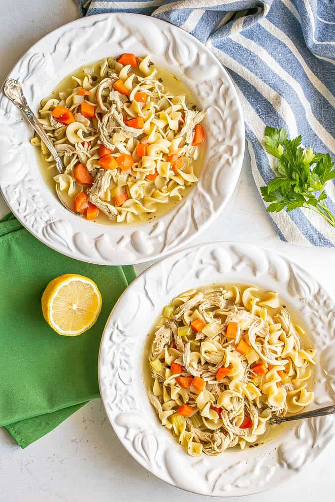 Two pretty white bowls of chicken noodle soup with spoons resting in them, and green napkins nearby along with a halved lemon and some parsley sprigs