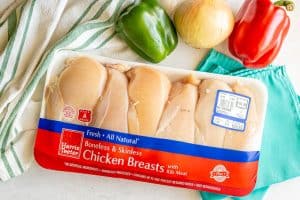 A large pack of chicken breasts with fresh peppers and an onion nearby