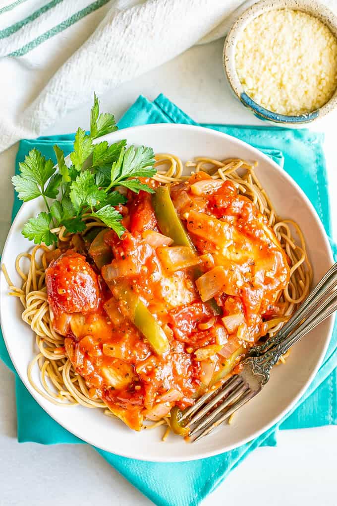 Chicken cacciatore served over spaghetti noodles in a shallow white bowl with parsley to the side and Parmesan cheese in a bowl nearby