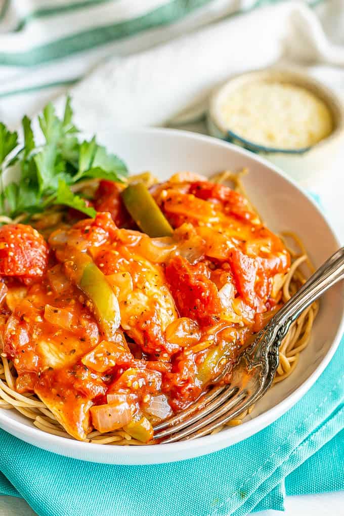 Chicken cacciatore served over spaghetti noodles in a shallow white bowl with parsley and Parmesan cheese to the side