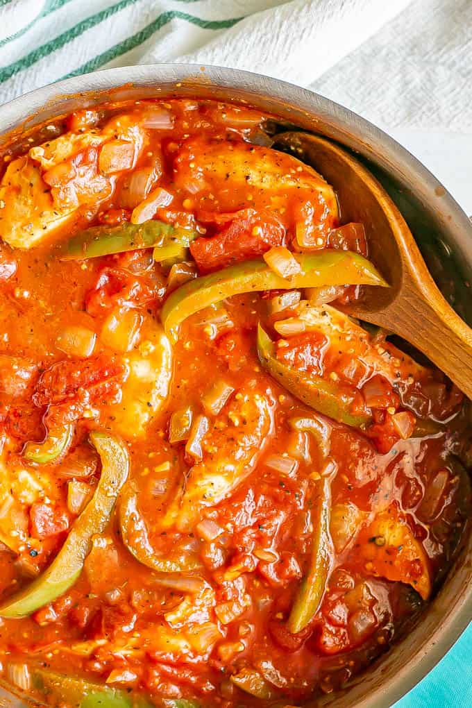 A large skillet filled with chicken strips, peppers and tomato sauce with a spoon resting in it