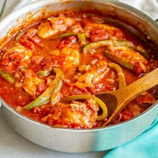 A wooden spoon lifting up a piece of chicken cacciatore from a large skillet