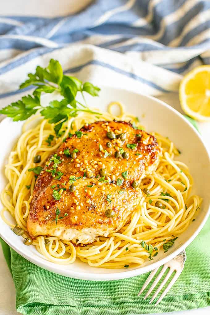 Chicken piccata served over thin spaghetti noodles in a low shallow white bowl with parsley sprinkled on top