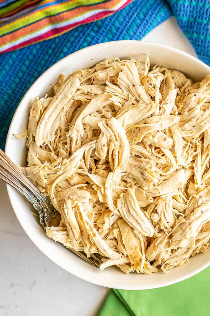 A white bowl full of shredded chicken cooked in a slow cooker with two forks tucked in the bowl