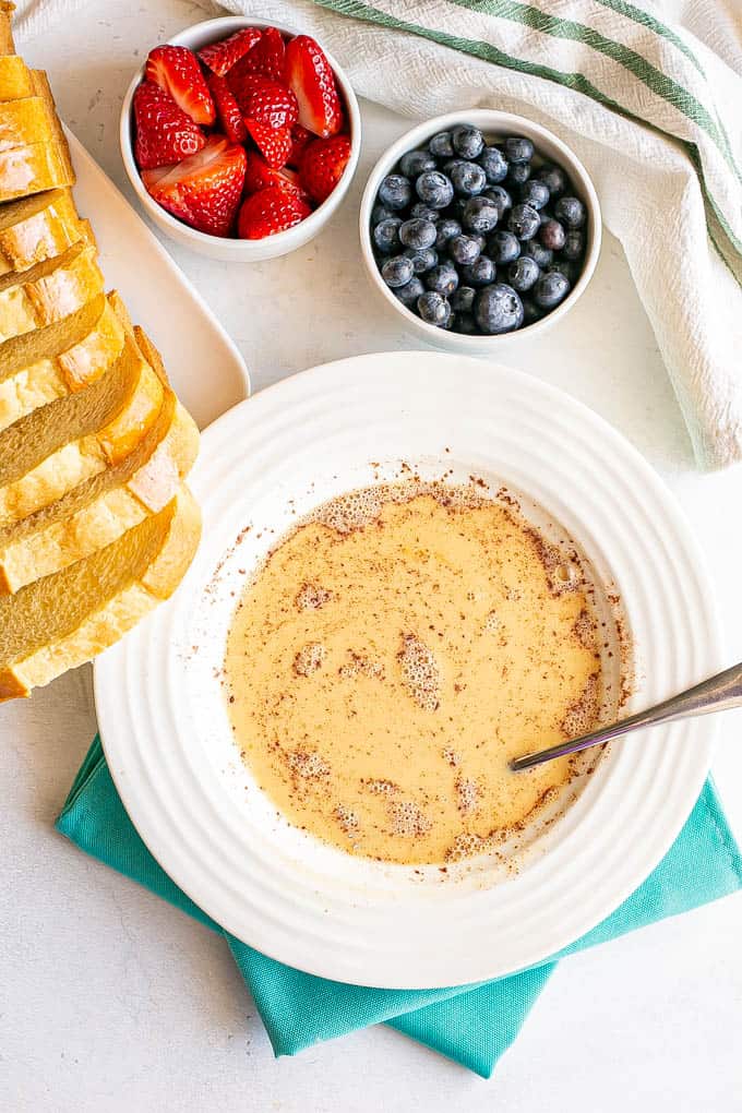 A bowl of an eggy mixture for French toast surrounded by fresh berries and sliced brioche bread