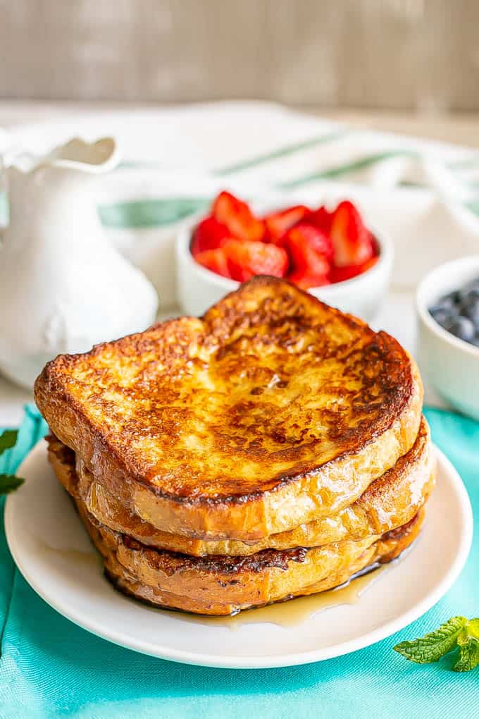 A small white plate with three stacked slices of French toast drizzled with maple syrup with bowls of fresh berries nearby