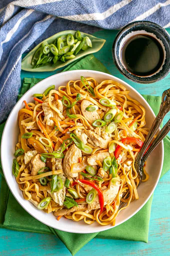 Overhead shot of chicken lo mein served in a shallow white bowl with two forks resting in the bowl and soy sauce and green onions to the side