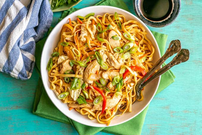 Overhead shot of chicken lo mein served in a shallow white bowl with two forks resting in the bowl and soy sauce to the side