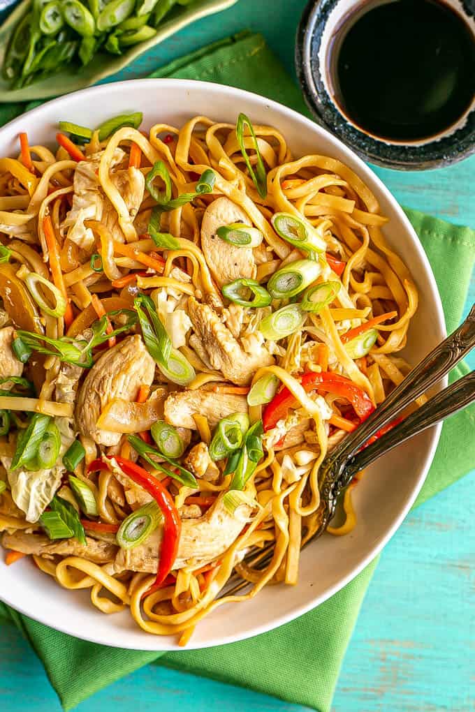 Overhead shot of chicken lo mein served in a shallow white bowl with two forks resting in the bowl and soy sauce to the side