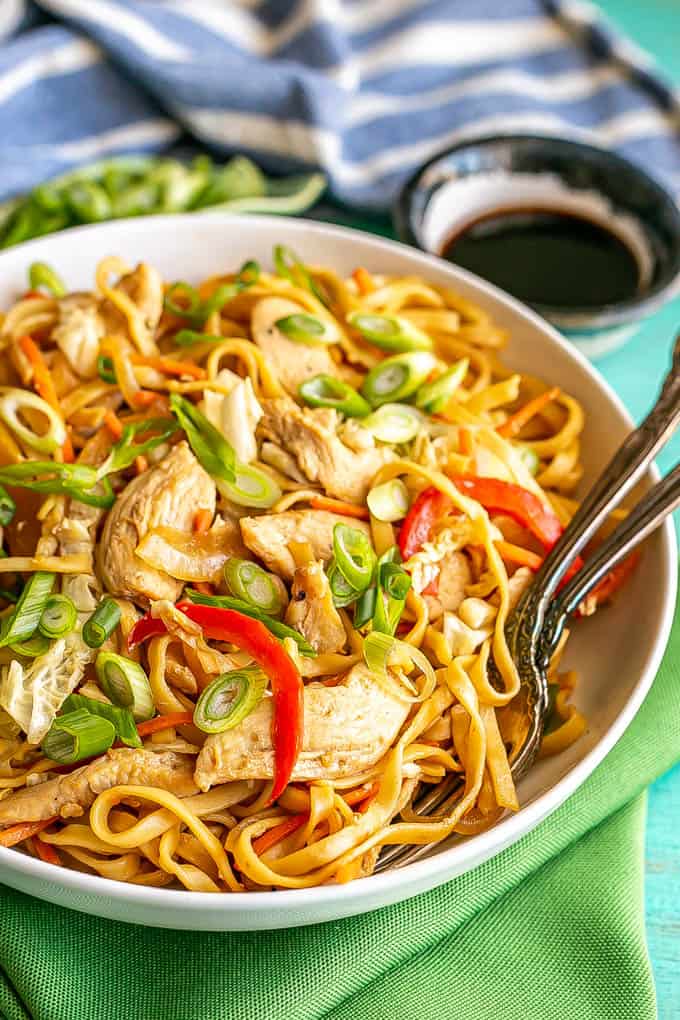 Close up of a shallow, wide white bowl full of chicken lo mein topped with sliced green onions with two forks resting in the bowl