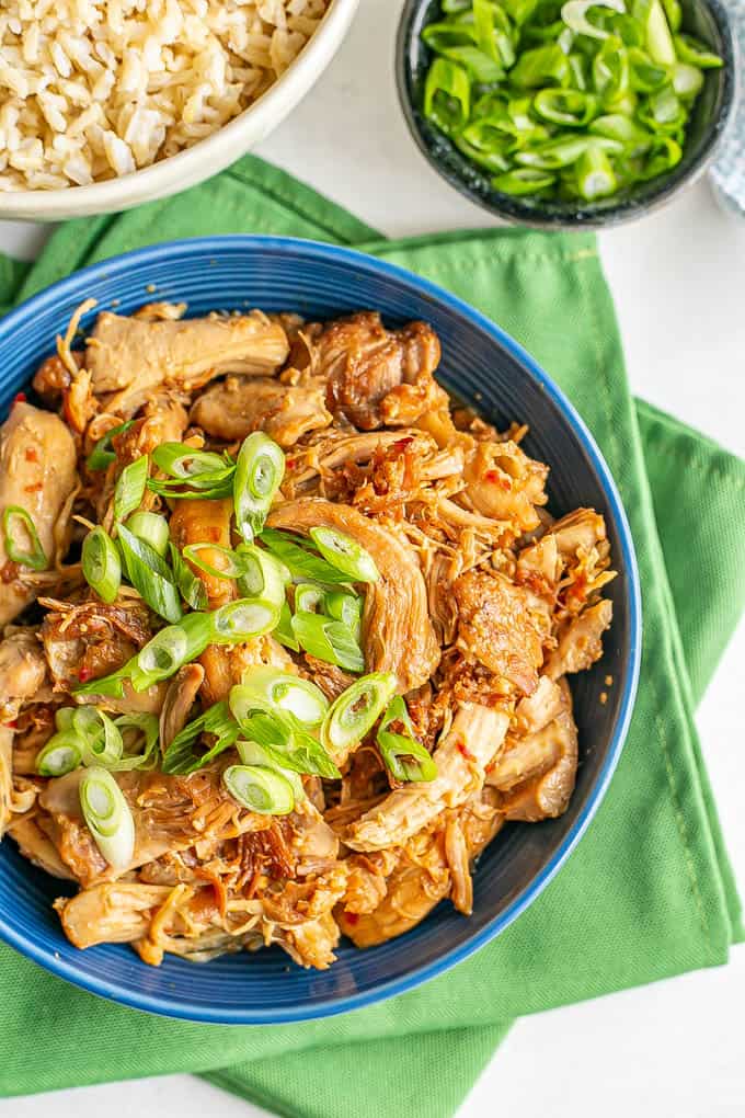 Close up of honey garlic chicken in a blue serving bowl topped with green onions with a bowl of rice to the back