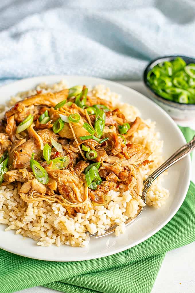 A white plate set on green napkins with brown rice topped with shredded honey garlic chicken and green onions and a fork alongside