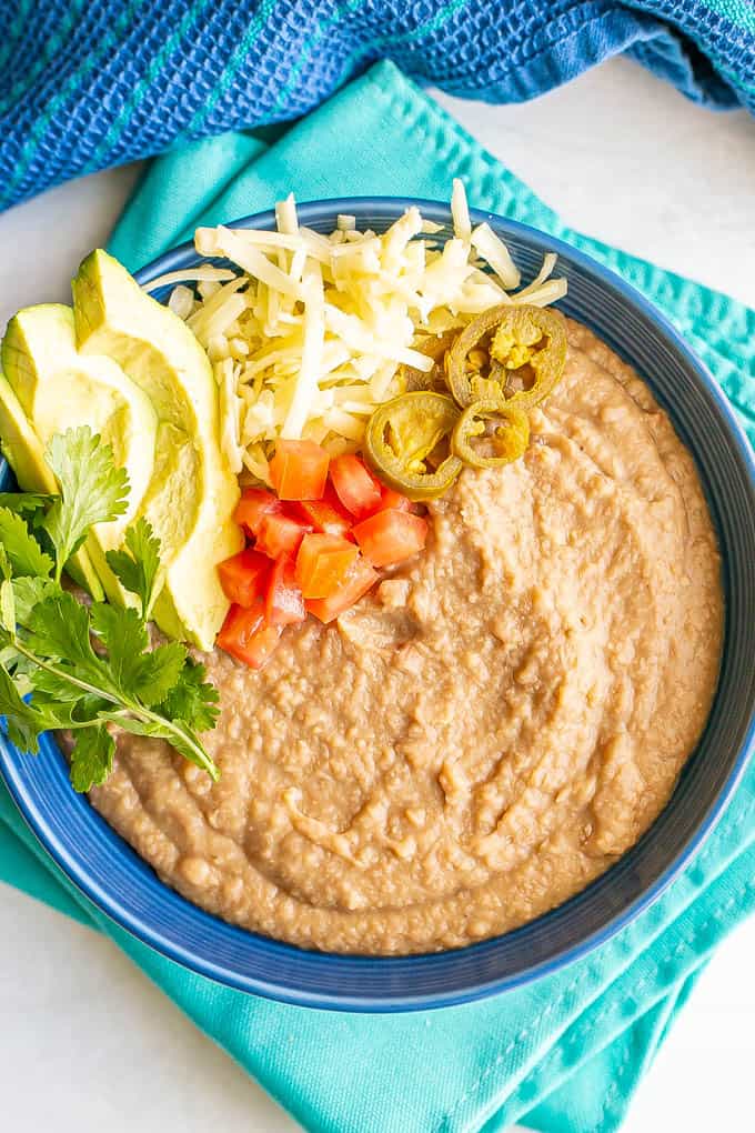 A blue bowl with homemade refried beans topped with cheese, tomatoes, avocado, cilantro and pickled jalapenos