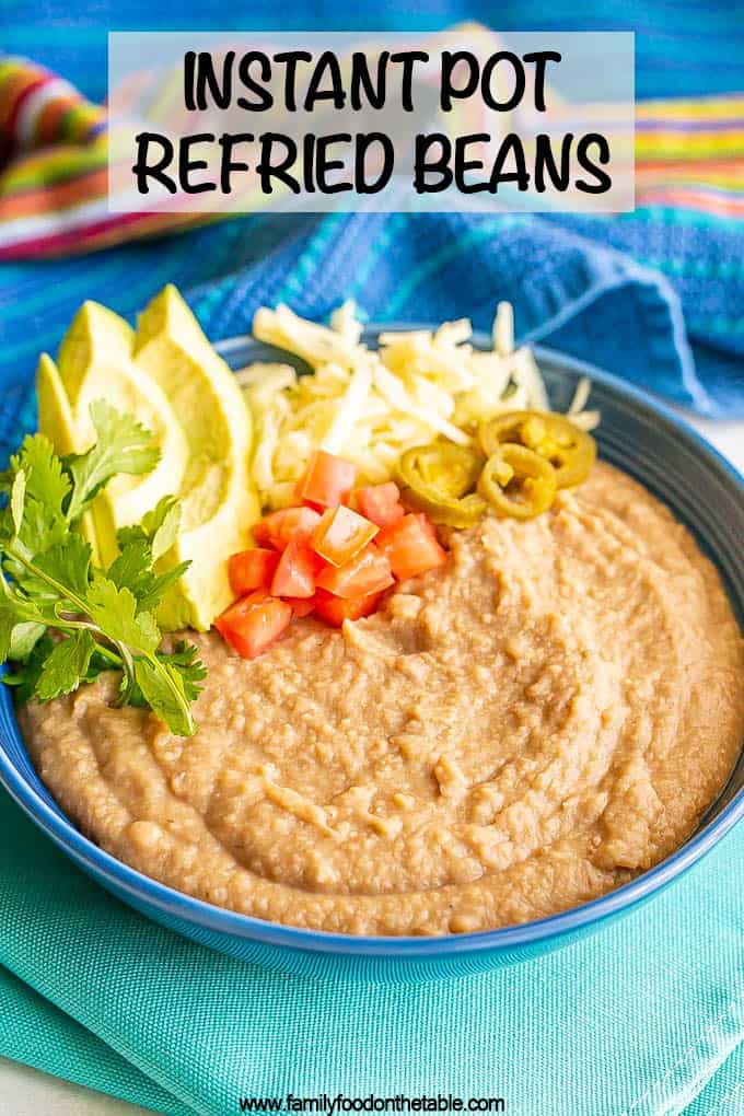 Creamy homemade refried beans in a blue bowl with cilantro, avocado, tomatoes, cheese and pickled jalapenos to the side of the bowl as toppings and a text overlay on the photo