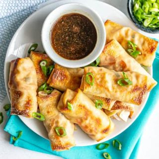 A plate of stacked baked vegetable egg rolls with a small bowl of dipping sauce and green onions sprinkled on top