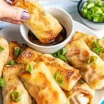 A hand dipping a baked vegetable egg roll into a soy sauce based dipping sauce with a text overlay on the photo