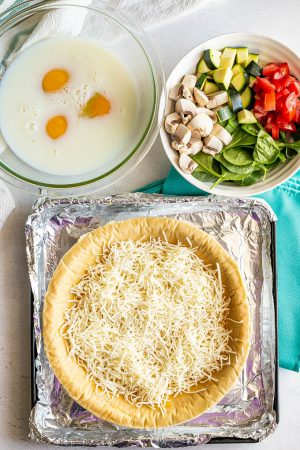 A pie crust sprinkled with cheese alongside a bowl of fresh vegetables and a glass bowl with milk and eggs