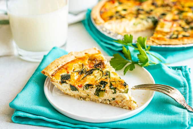 A slice of veggie loaded quiche served on a small white plate set on some turquoise napkins