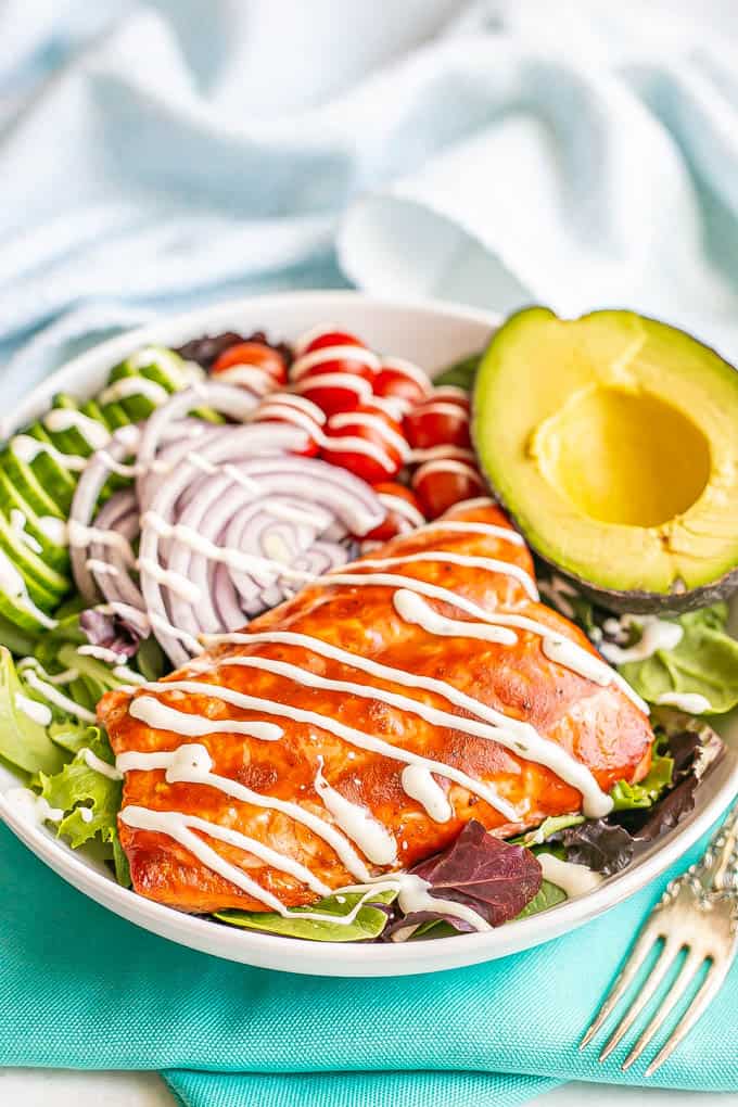 BBQ salmon atop a spring greens and veggie salad in a large white bowl with a half an avocado to the side