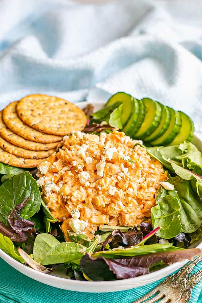 A mound of buffalo chicken salad atop mixed greens in a salad bowl with cucumber slices and whole grain crackers