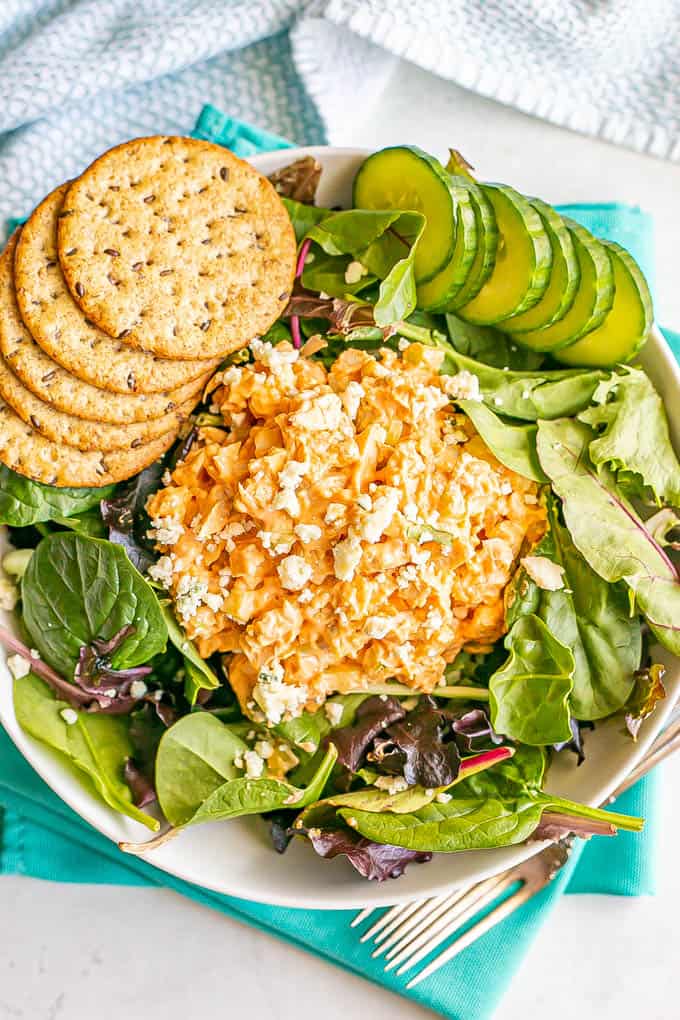 Buffalo chicken salad with blue cheese crumbles served on mixed greens in a large salad bowl with cucumber and crackers to the side