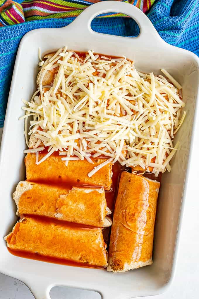 A white baking dish with chicken enchiladas being assembled and topped with cheese before baking