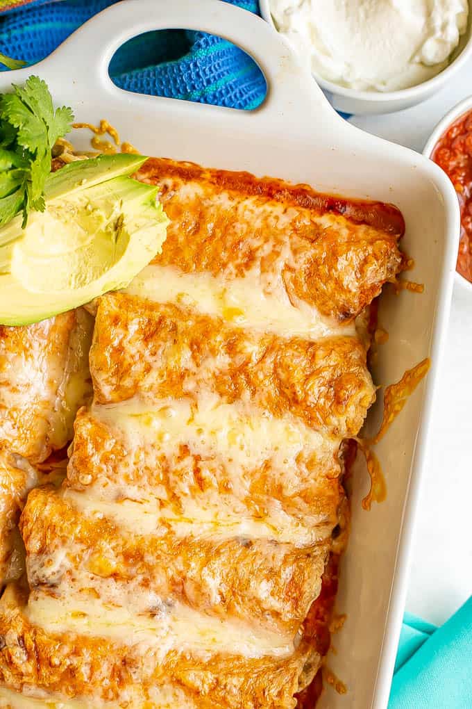 A white pan filled with cheesy chicken enchiladas with avocado slices to the side