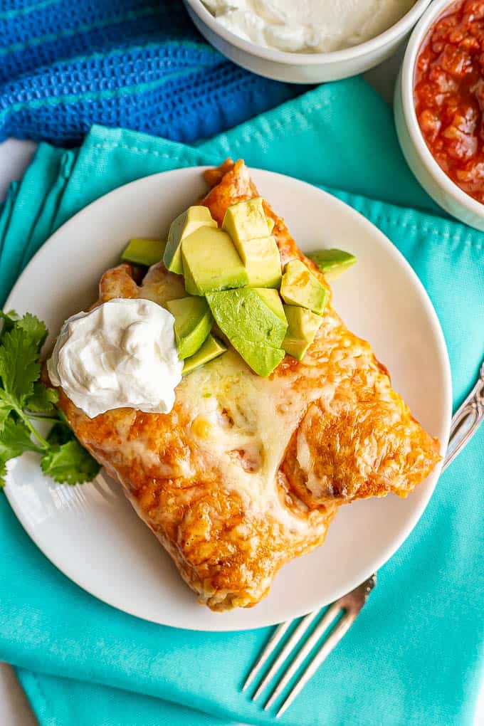 Two cheesy chicken enchiladas served on a small white plate with sour cream, avocado and cilantro on top