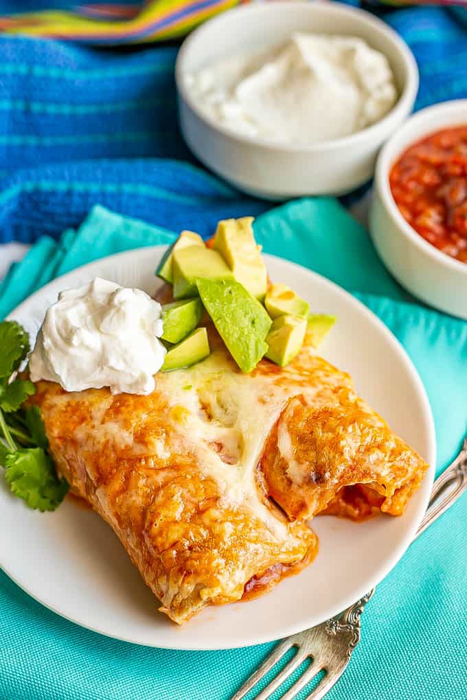 Two cheesy chicken enchiladas served on a small white plate with sour cream, avocado and cilantro on top and salsa and sour cream in bowls in the background