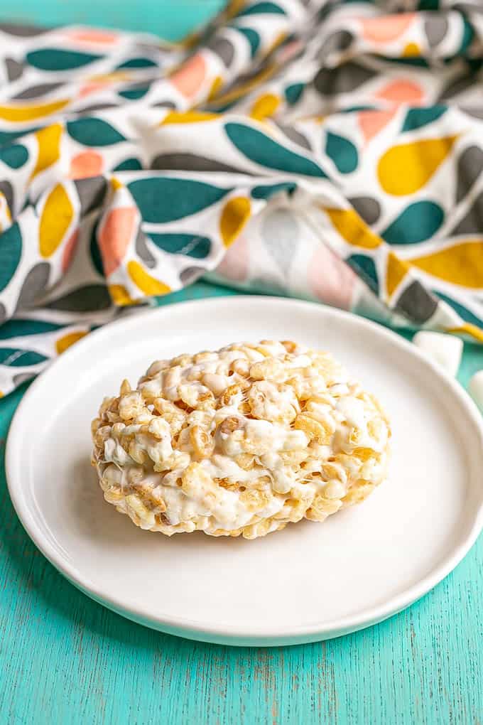 A single Rice Krispie treat on a small white plate with a colorful hand towel in the background