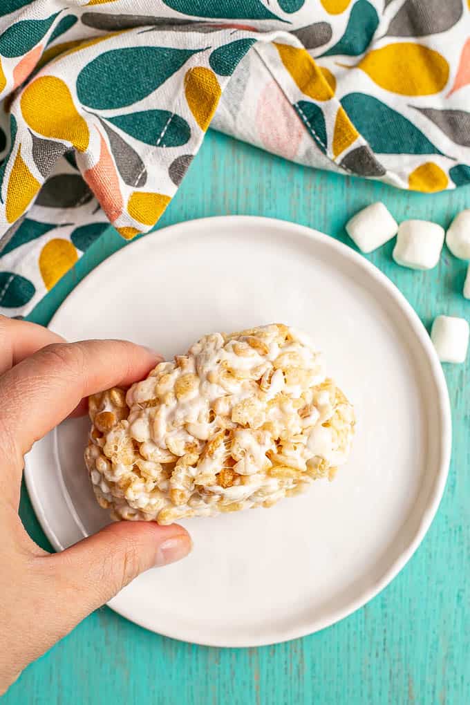 A hand holding a gooey Rice Krispie treat on a small white plate with mini marshmallows scattered nearby